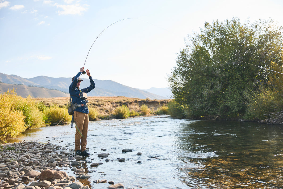 A fly fisherman wearing Steely Zips famous Rocky Mountain Grid Fleece stands in the BigWood river reeling in a trout fish with mountains and green trees in the background.