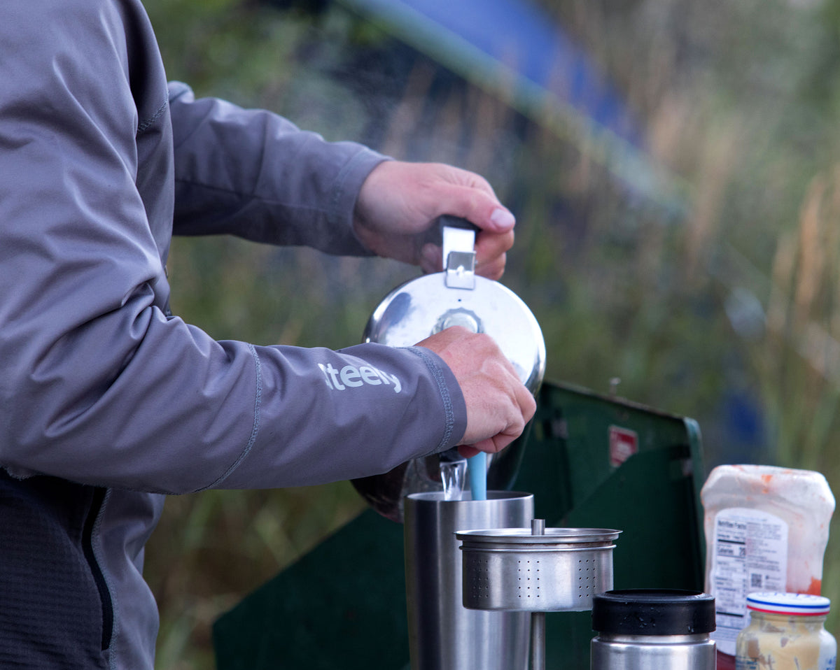 A fisherman on a camping trip wearing a gray Steely Zips Hoodie jacket pours coffee from a stainless steel kettle into a metal travel mug outdoors.