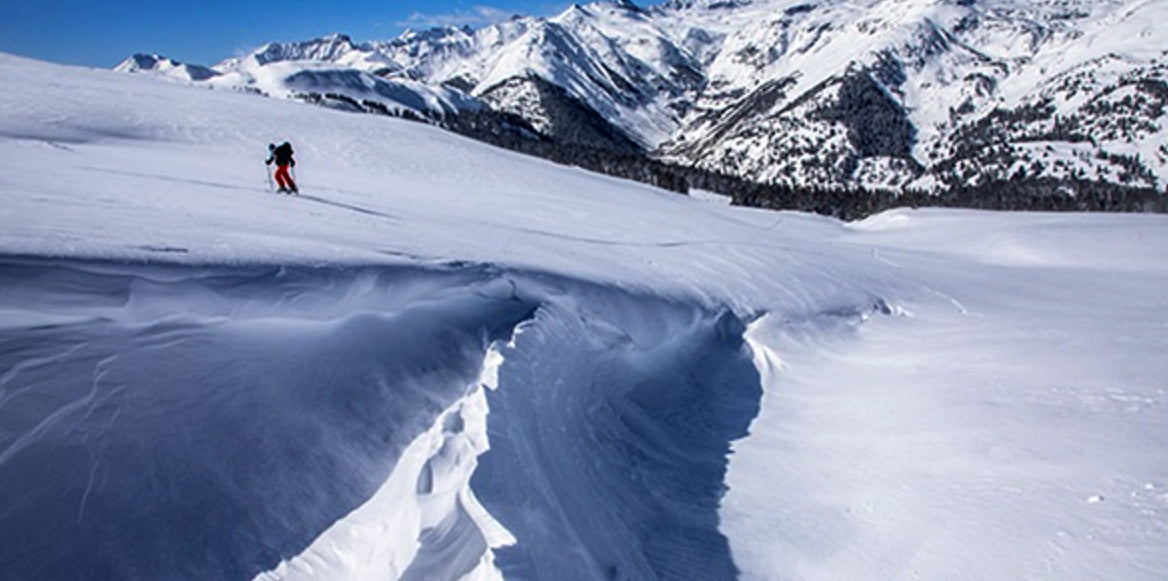 A snowy mountain landscape with a  backcountry skier hiking on skies, with jagged snow-covered peaks and blue sky in the background.