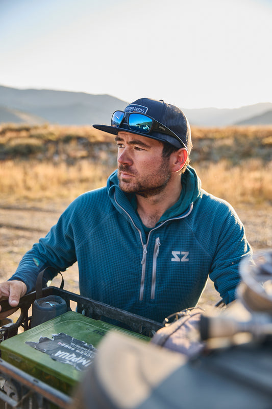 A man wearing a Ocean Blue Rocky Mountain Grid Fleece hoodie, a dark blue cap with sunglasses, and a green fly-box sits on an all-terrain vehicle in an outdoor desert landscape.