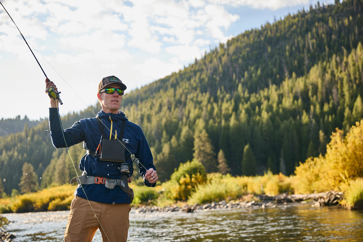 A man wearing Steely Zips Rocky Mountain Grid Fleece fly fishing in the Big Wood River in Idaho. He is also wearing  tan pants, sunglasses, and a cap, with a pine tree mountains in the background.