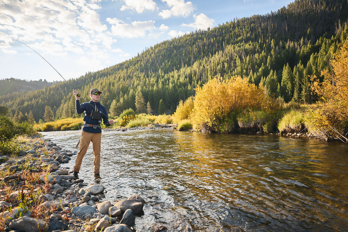 A man fly fishing in Steely Zips Rocky Mountain Grid Fleece, standing on stones in the water and casting a rod. He is wearing a dark blue Steely Zips Hoodie, tan pants, and sunglasses.