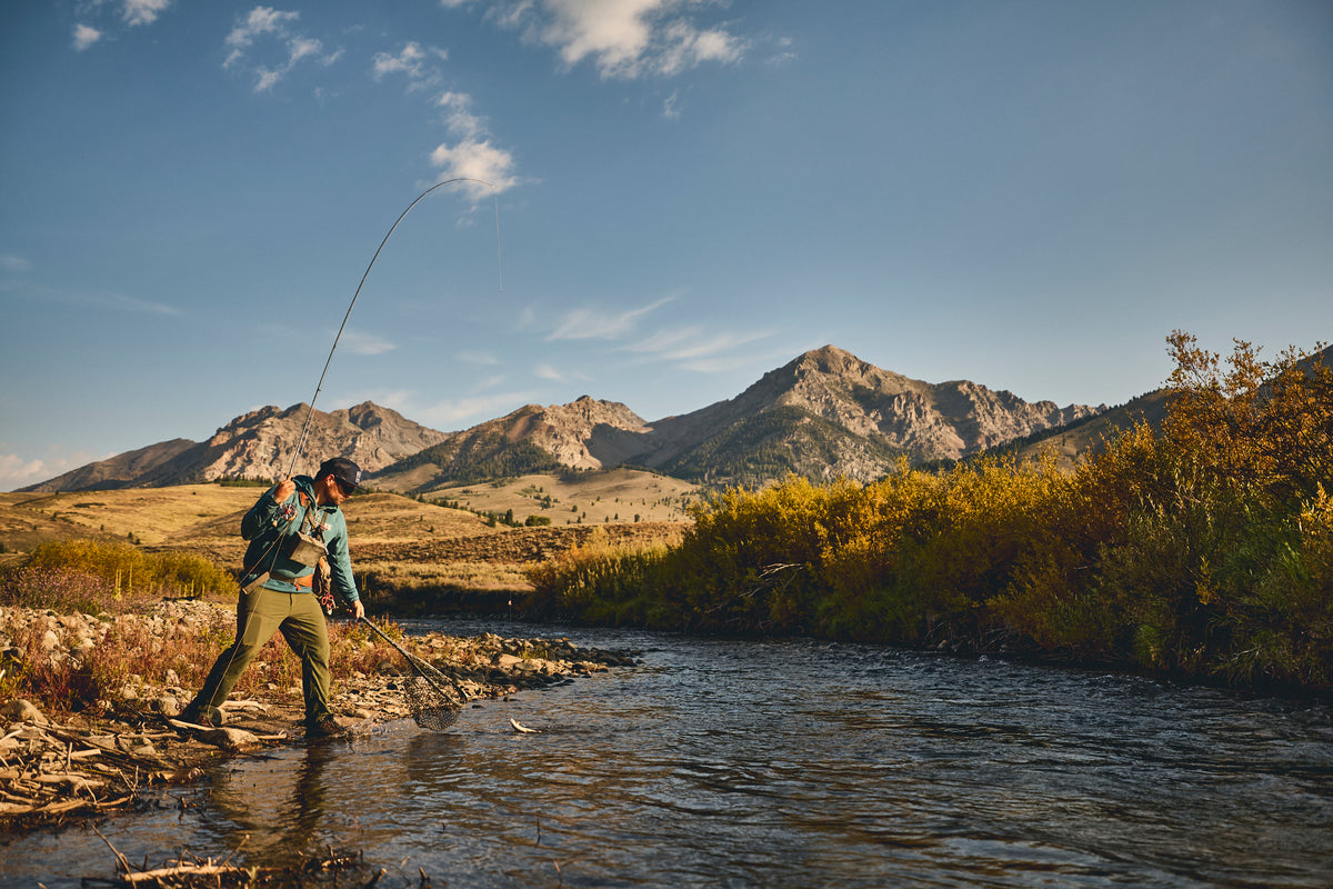 A fly fisherman wearing a Steely Zips Grid Fleece stands in a river, casting a rod with a mountain range and autumn trees in the background.