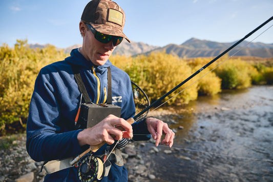 A man wearing a blue Steely Zips Rocky Mountain grid fleece hoodie, sunglasses, and a camouflage cap is fly fishing in the BIg Wood river with mountains in the background.