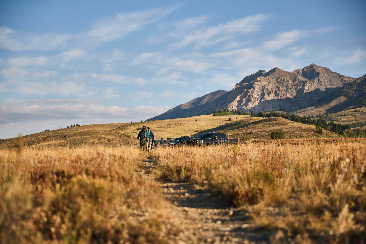 A scenic outdoor landscape with a dirt path through dry golden grass leading toward a mountain range under a blue sky with wispy clouds. Two fisherman wearing Steely Zips Rocky Mountain Grid Fleeces are walking on a dirt trail to the Big Wood River. 